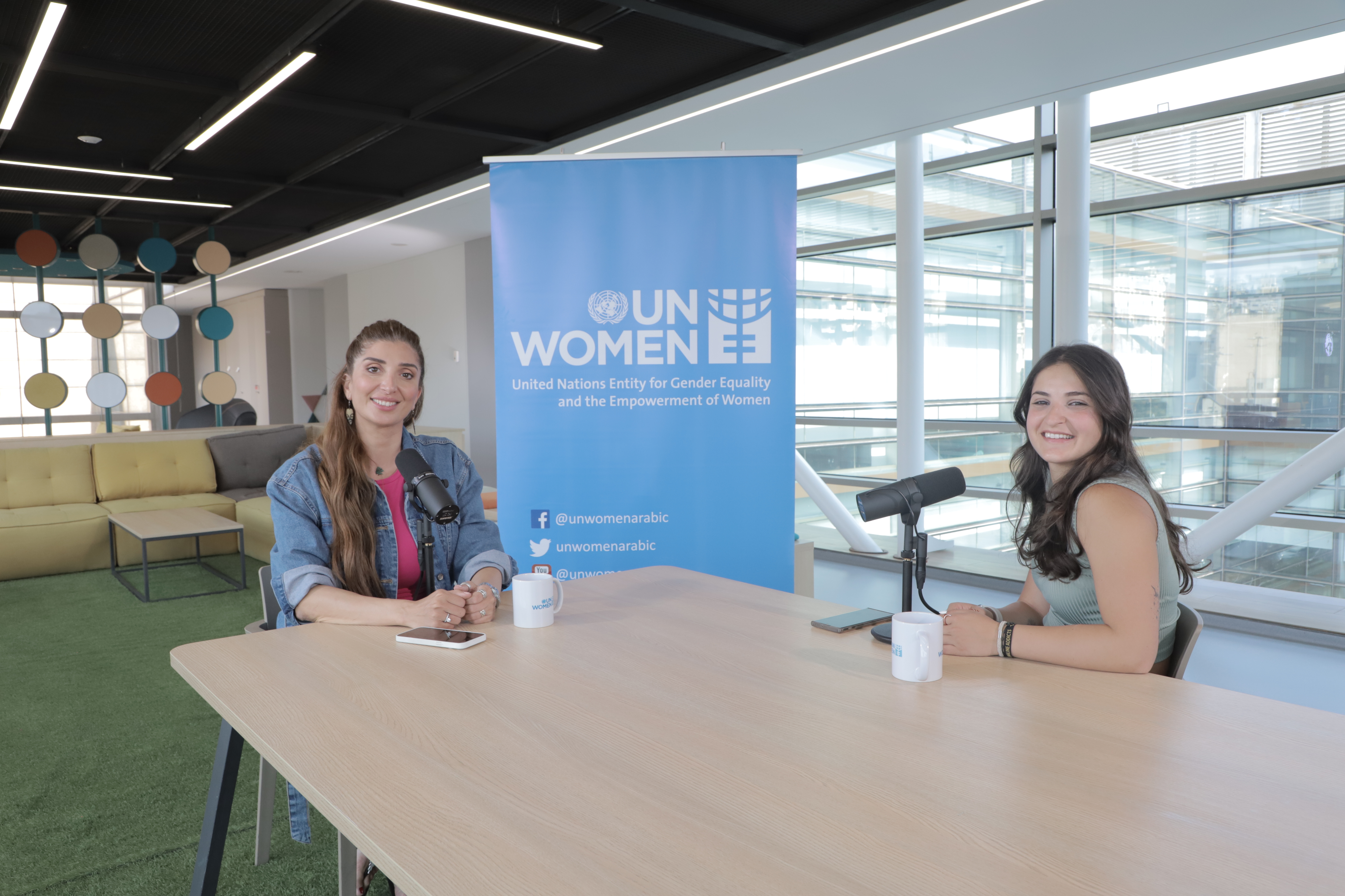 Two young women facing the camera with microphones in front of them. UN Women banner in the back.